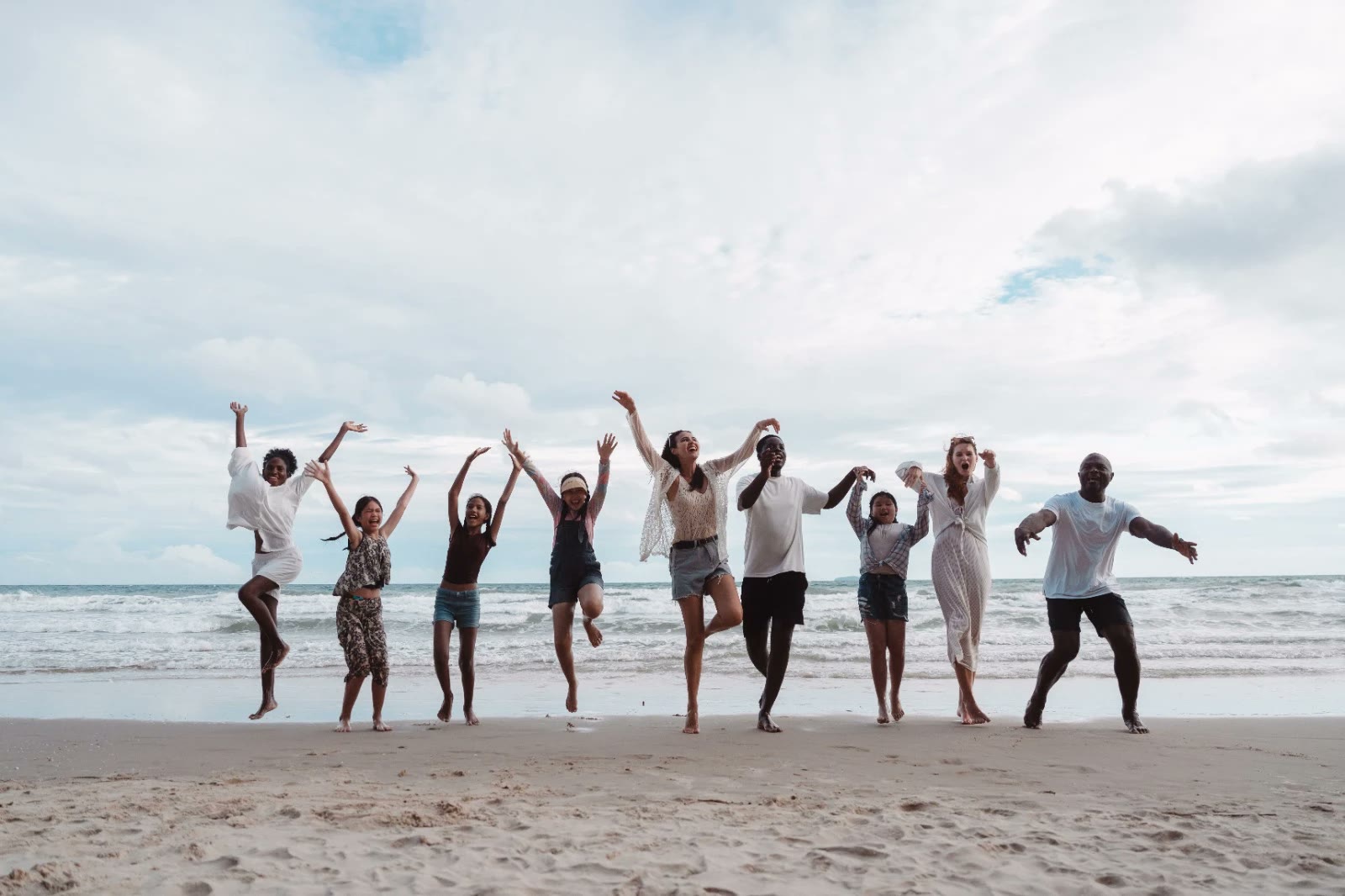 A group of friends leap and laugh with arms raised on a sandy beach at the water's edge.