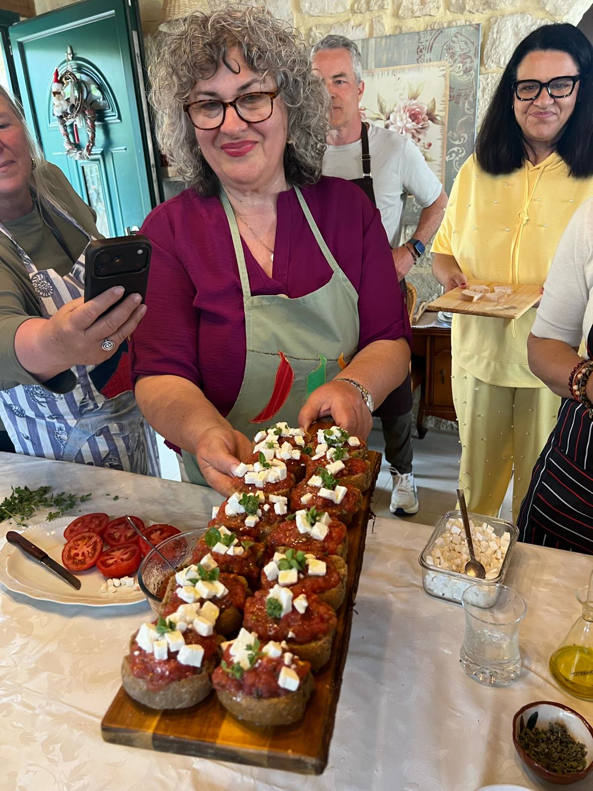 A local host presents a board of dakos topped with tomato, feta and herbs while guests look on.