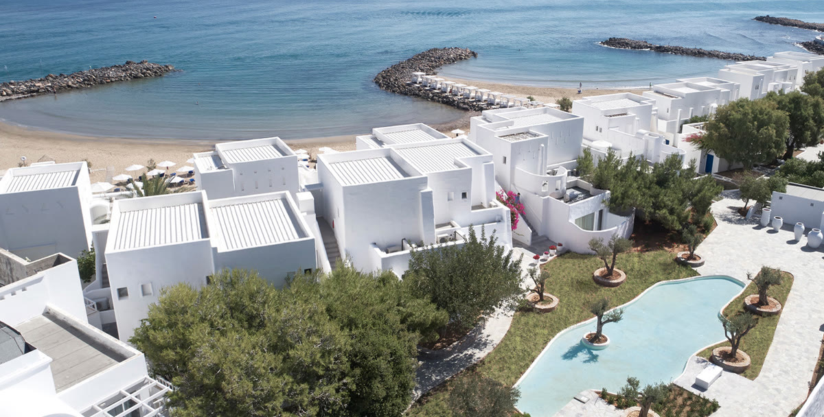 Aerial view of Knossos Beach Resort: white cubic buildings, olive trees and a pool beside a sandy Mediterranean beach.