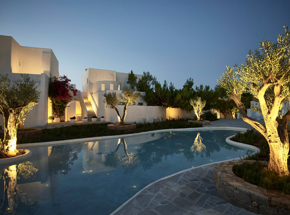 At dusk, uplit olive trees and a dry-stone planter wall beside a curving pool between whitewashed buildings.