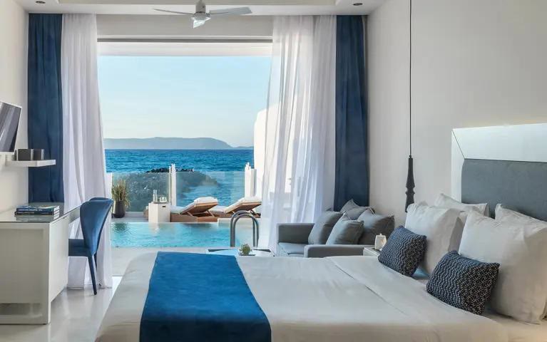 A white and blue guest room opening through sheer curtains onto a pool and the sea.