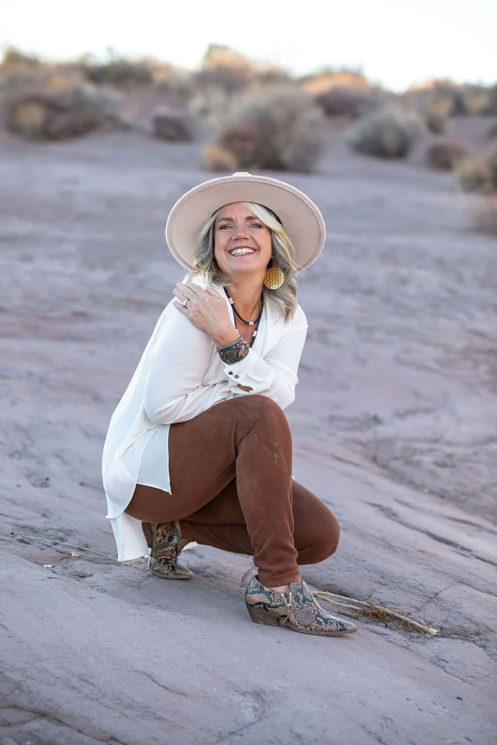 Wendy Bunnell, smiling in a wide-brimmed hat and white shirt, crouched on sunlit sandstone.